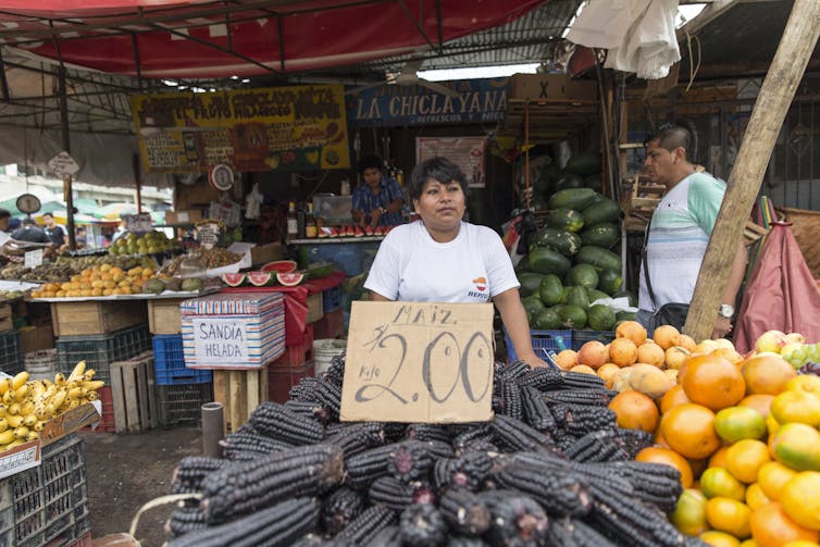 Uma pessoa fica atrás de uma mesa cheia de espigas de milho roxas em um mercado de agricultores.